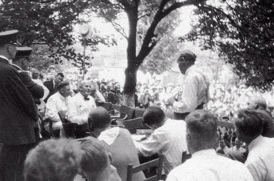 'The duel in the Shade.'  Bryan is seated center left, Darrow on the right. Most of the audience are in bright sunsghine beyond Bryan and Darrow.